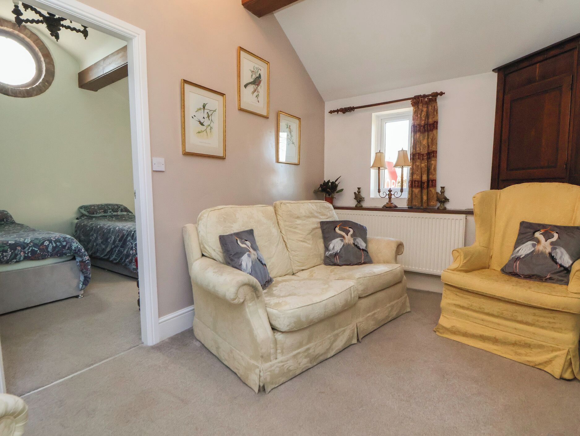 A cozy living room with a beige sofa, a yellow armchair, and two matching armchairs. There are three framed bird paintings on the wall, a window with curtains, and a radiator.