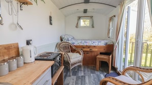 Una cucina compatta con un piano cottura, un bollitore e contenitori per la conservazione. Un letto con una copripiumino floreale si trova accanto alla cucina, e una sedia in vimini è posizionata vicino alla finestra.