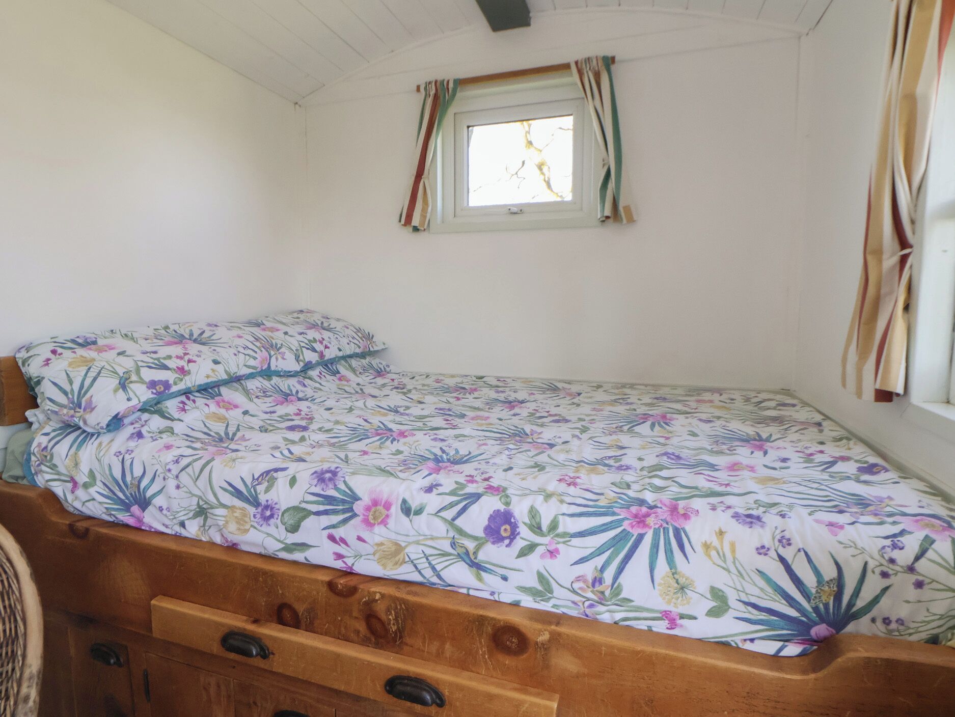 Pequeña habitación con cama individual, colcha de flores, cabecera de madera, ventana con cortinas y pared blanca.