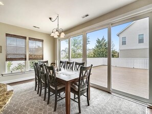 A dining area with a table and chairs, large windows, and a view of a house and deck. - Modern 4BR House near Bethany Beach | Decks & Pool (Bethany Beach)