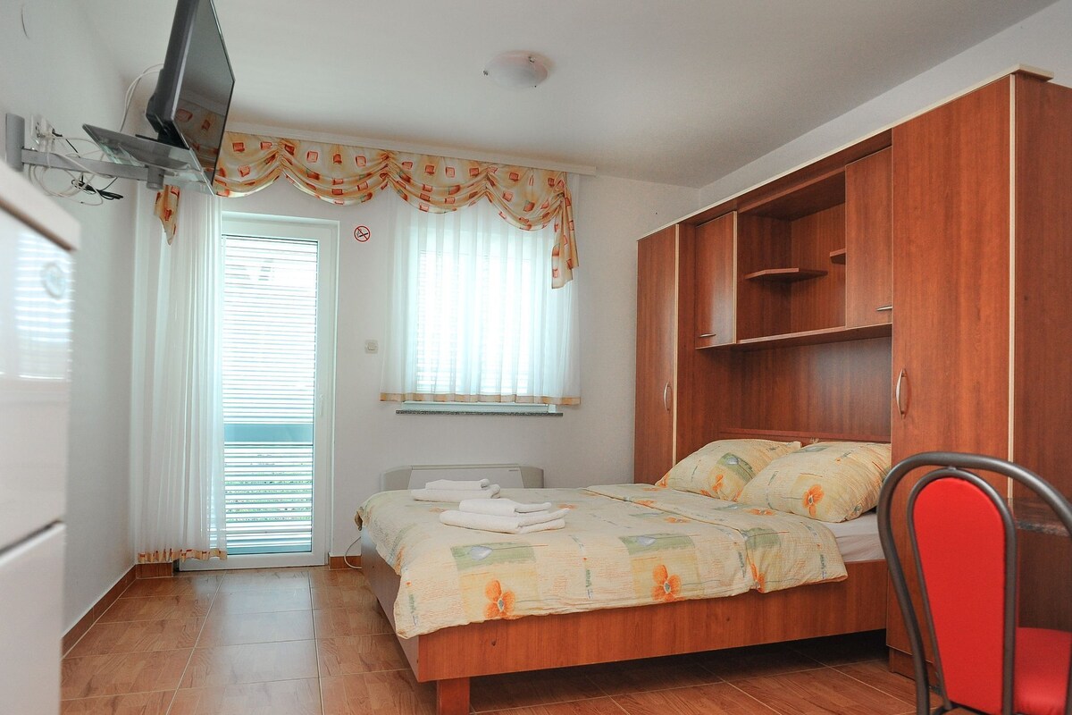 A bedroom with a bed, a wooden wardrobe, a TV mounted on the wall, and a window with curtains.