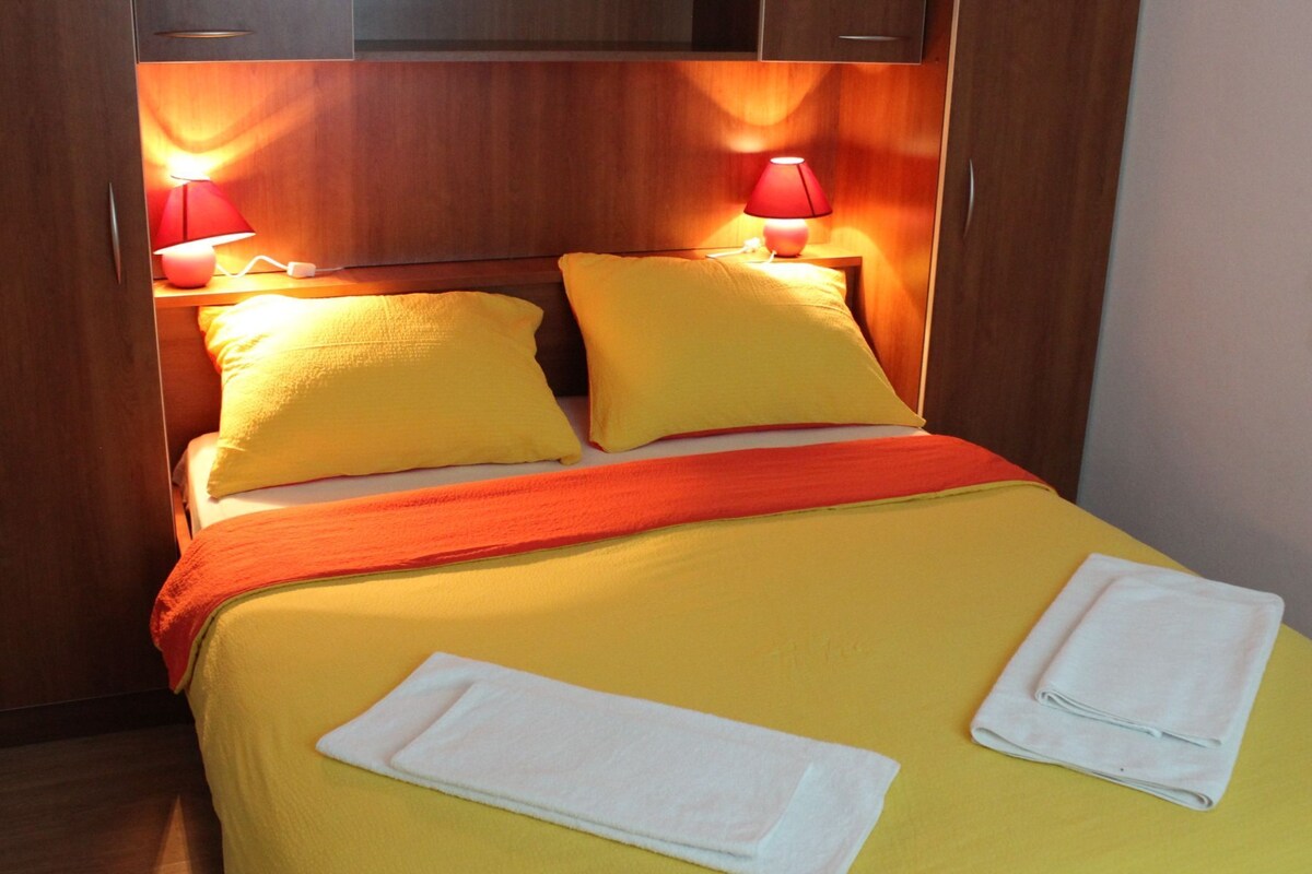 A bed with yellow bedding, two pillows, a red blanket, and two white towels folded on the bed.