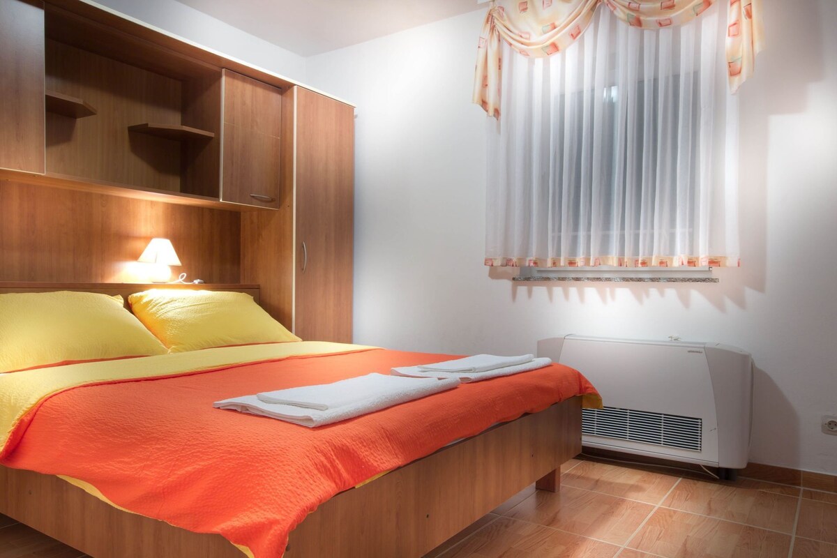 A hotel room with a bed, wooden wardrobe, air conditioning unit, and a window with sheer curtains.