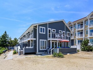 A two-story building with balconies, a covered outdoor seating area, and a pathway leading to the entrance. - Oceanfront 4BR Condo w/ Decks & BBQ | Sleeps 9! (Bethany Beach)
