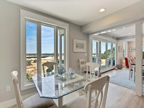 A modern dining area with a glass table and white chairs, large windows with a view of the beach, and a bright interior. - Oceanfront 4BR Condo w/ Decks & BBQ | Sleeps 9! (Bethany Beach)
