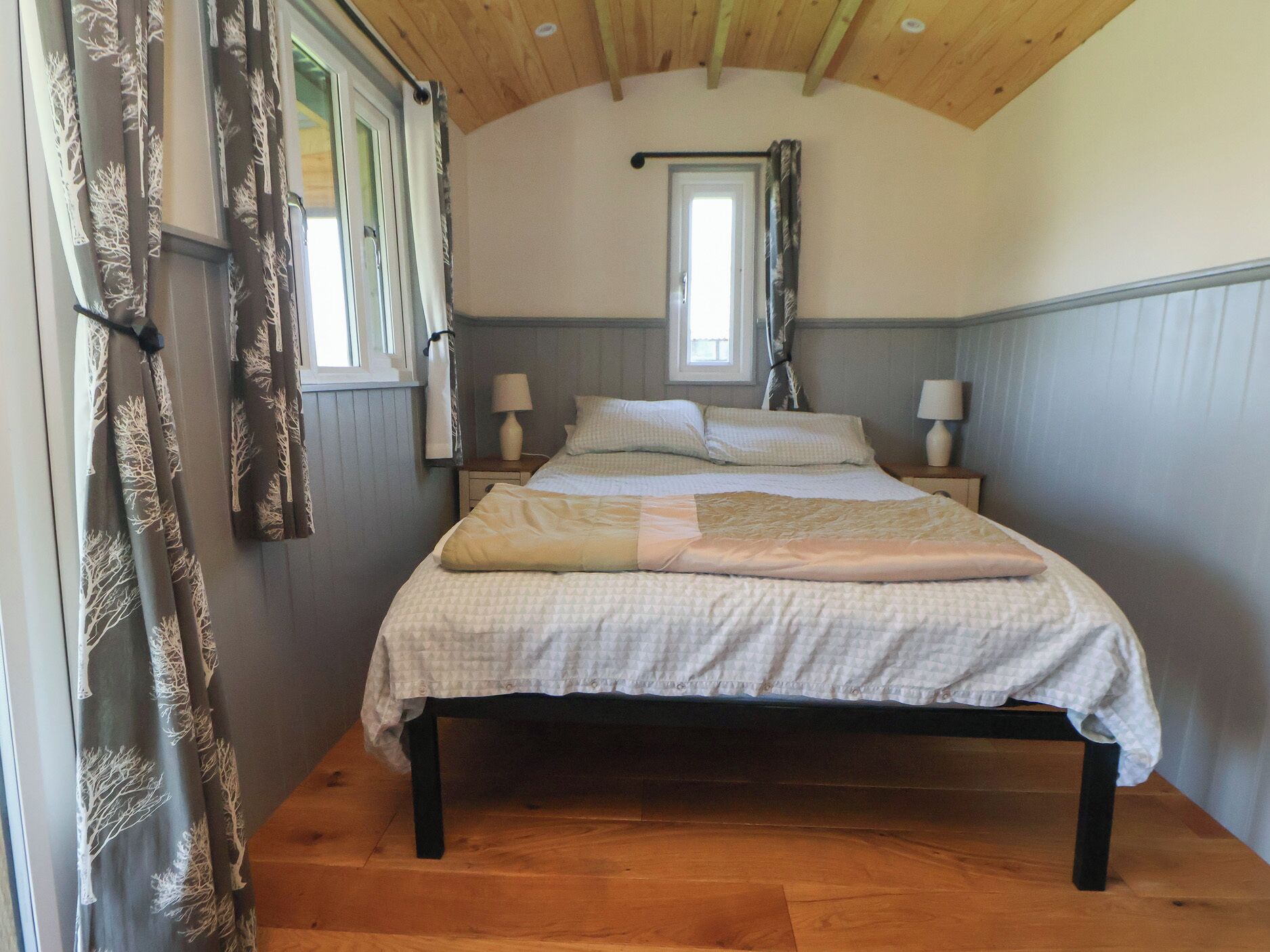 A bedroom with a wooden ceiling, a bed with a quilt, two bedside lamps, and curtains with a tree pattern.