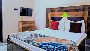 Un dormitorio con cabecera de madera, cama con varios cojines, mesita de noche con lámpara y televisión fijada a la pared.