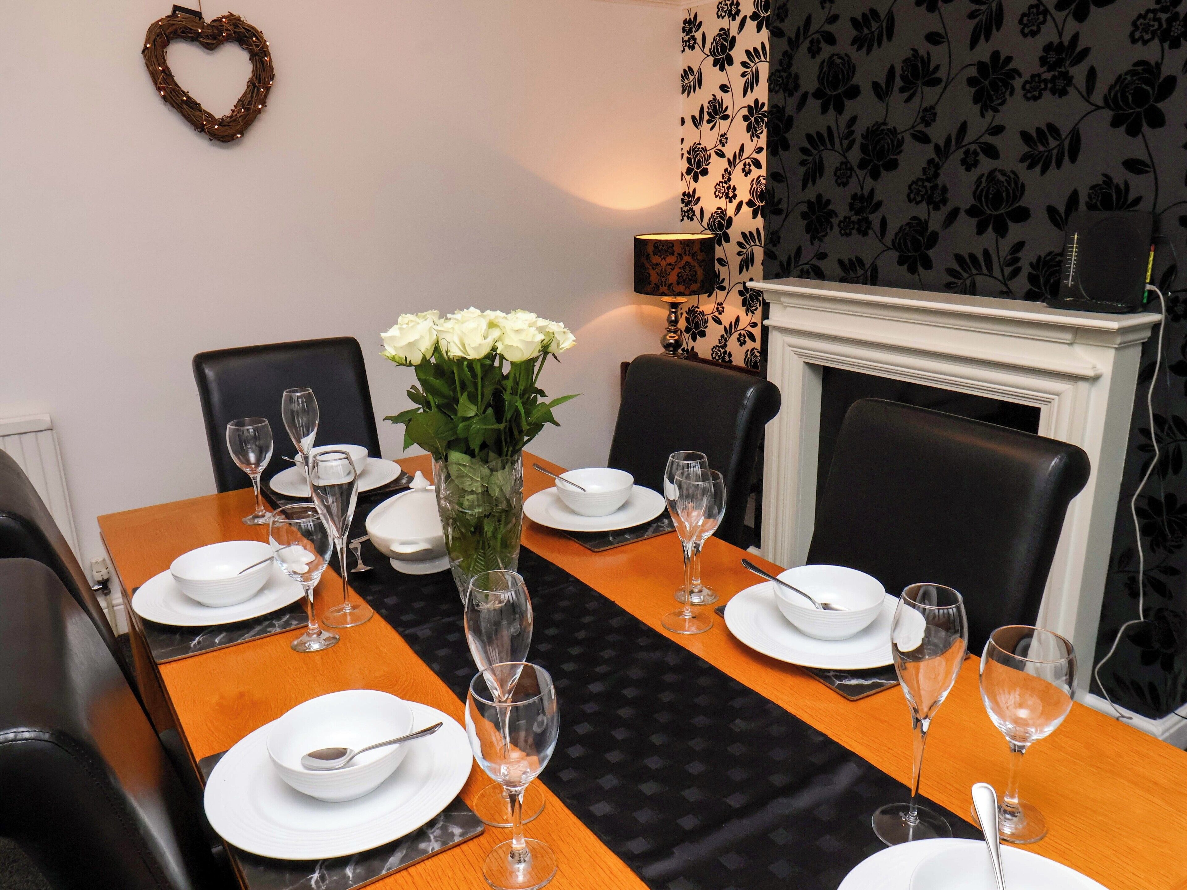 A dining room with a long table set for six, surrounded by black chairs, a vase of white flowers, and a fireplace.
