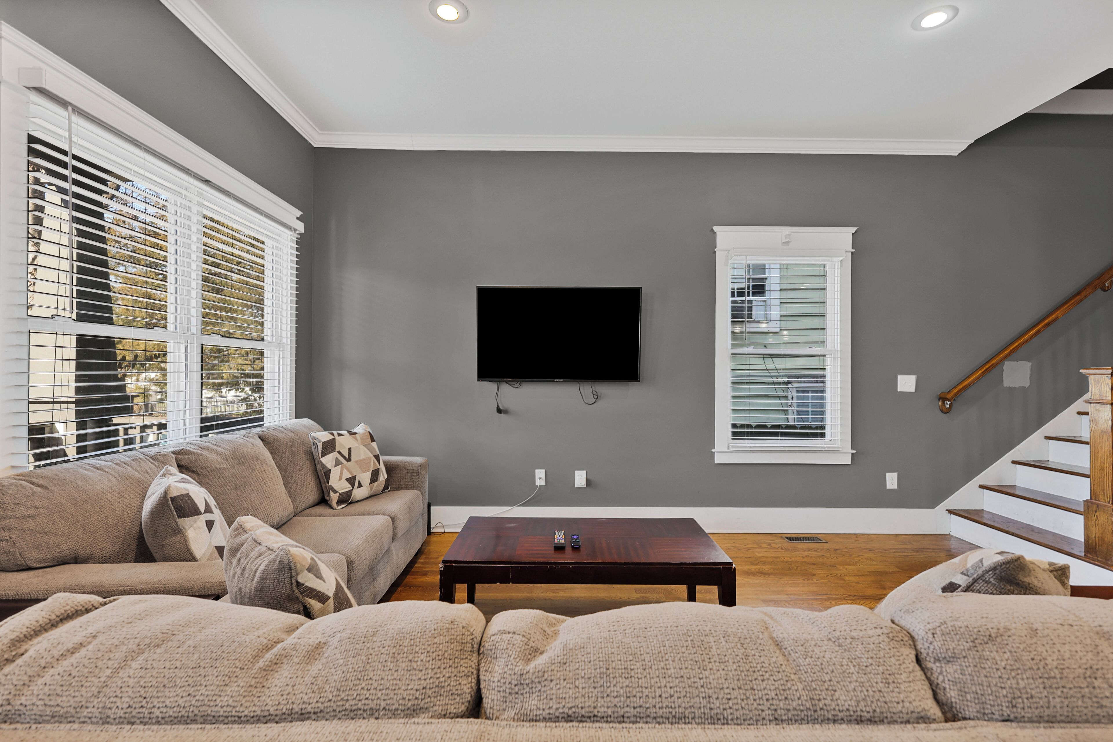 A living room with a gray wall, a flat-screen TV mounted above a wooden coffee table, and a staircase to the right.