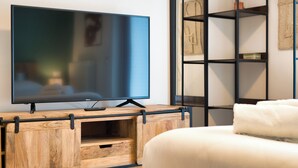 Een moderne woonkamer met een flatscreen televisie op een houten standaard, een witte bank en een zwarte boekenkast met diverse spullen.