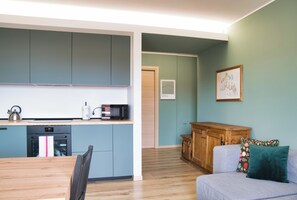 Ein kompakter Wohnbereich mit Küchenzeile, ausgestattet mit Mikrowelle, Wasserkocher und Backofen. Ein Holzschrank und ein graues Sofa mit einem Blumenkissen sind zu sehen.