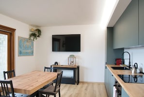 Eine moderne Küche mit einem Holzespeisetisch, Stühlen, einem über der Arbeitsfläche montierten Fernseher und einem kleinen Regal mit Pflanzen.