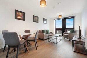 A modern living room with a glass dining table, two sofas, a TV on a wooden stand, and large windows. - Modern 1-Bed Apartment Near Liverpool City Centre (Merseyside)