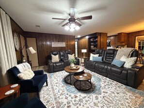A living room with a dark leather sofa, two armchairs, a coffee table, and a patterned rug. There is a ceiling fan, a clock, and a sign on the wall.