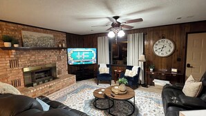 A living room with a brick fireplace, a television, a ceiling fan, a clock, and a large window with curtains.