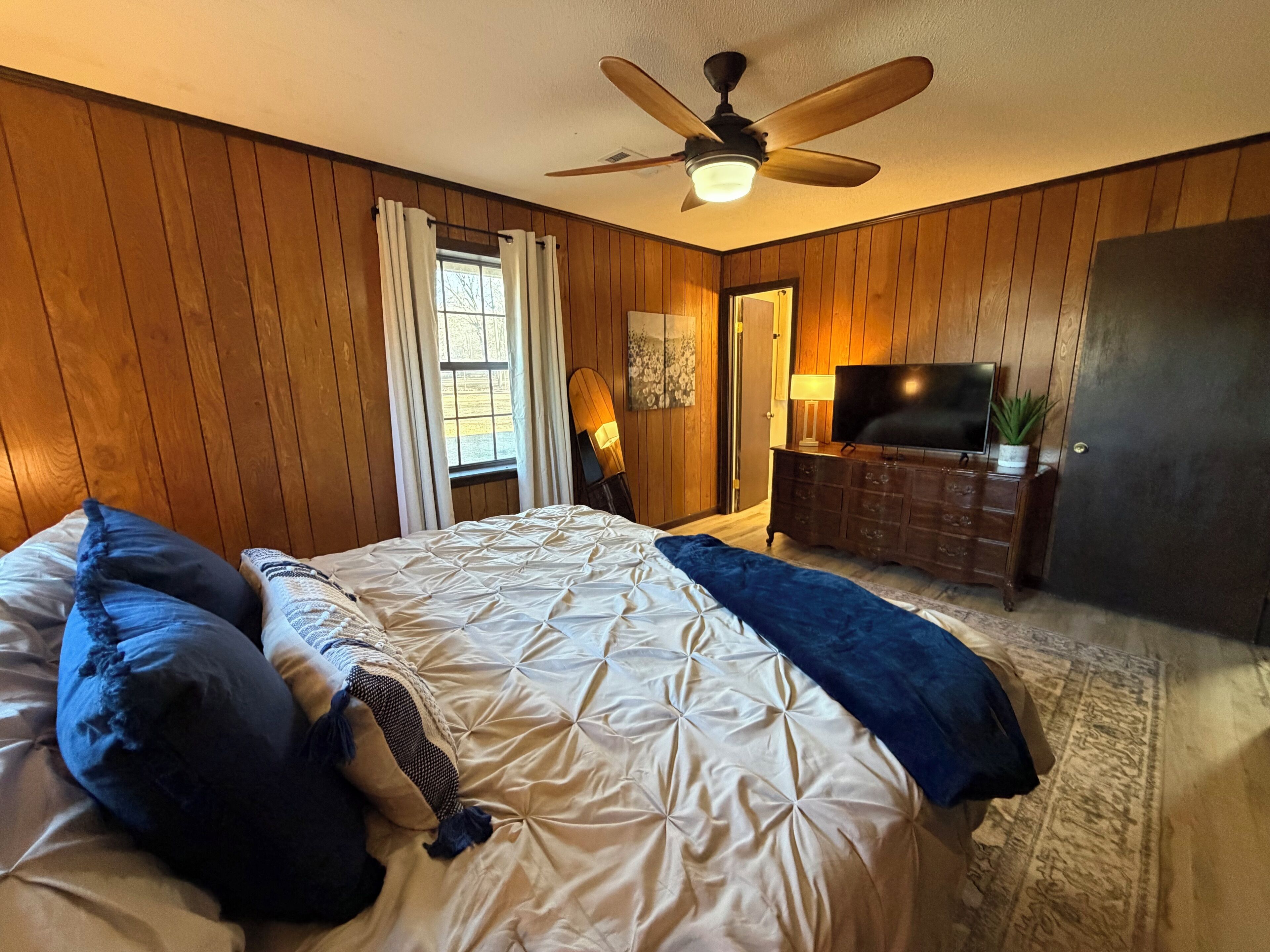 Une chambre à coucher avec un grand lit, des murs en bois, un ventilateur de plafond, une télévision et une porte donnant sur une autre pièce.