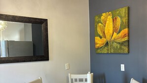 Spiseområde med et rundt bord, hvide stole, en lysekrone og et maleri af gule blomster på væggen.