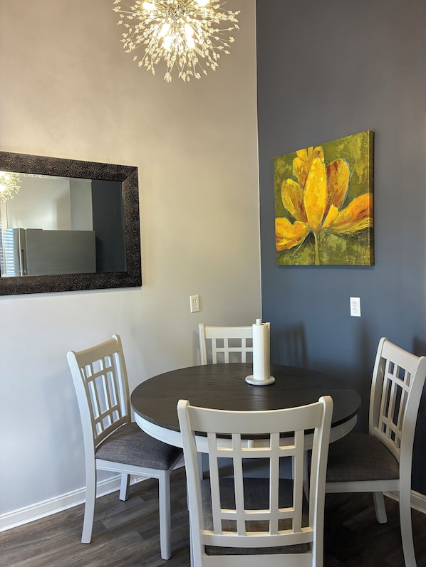 A dining area with a round table, white chairs, a chandelier, and a painting of yellow flowers on the wall.