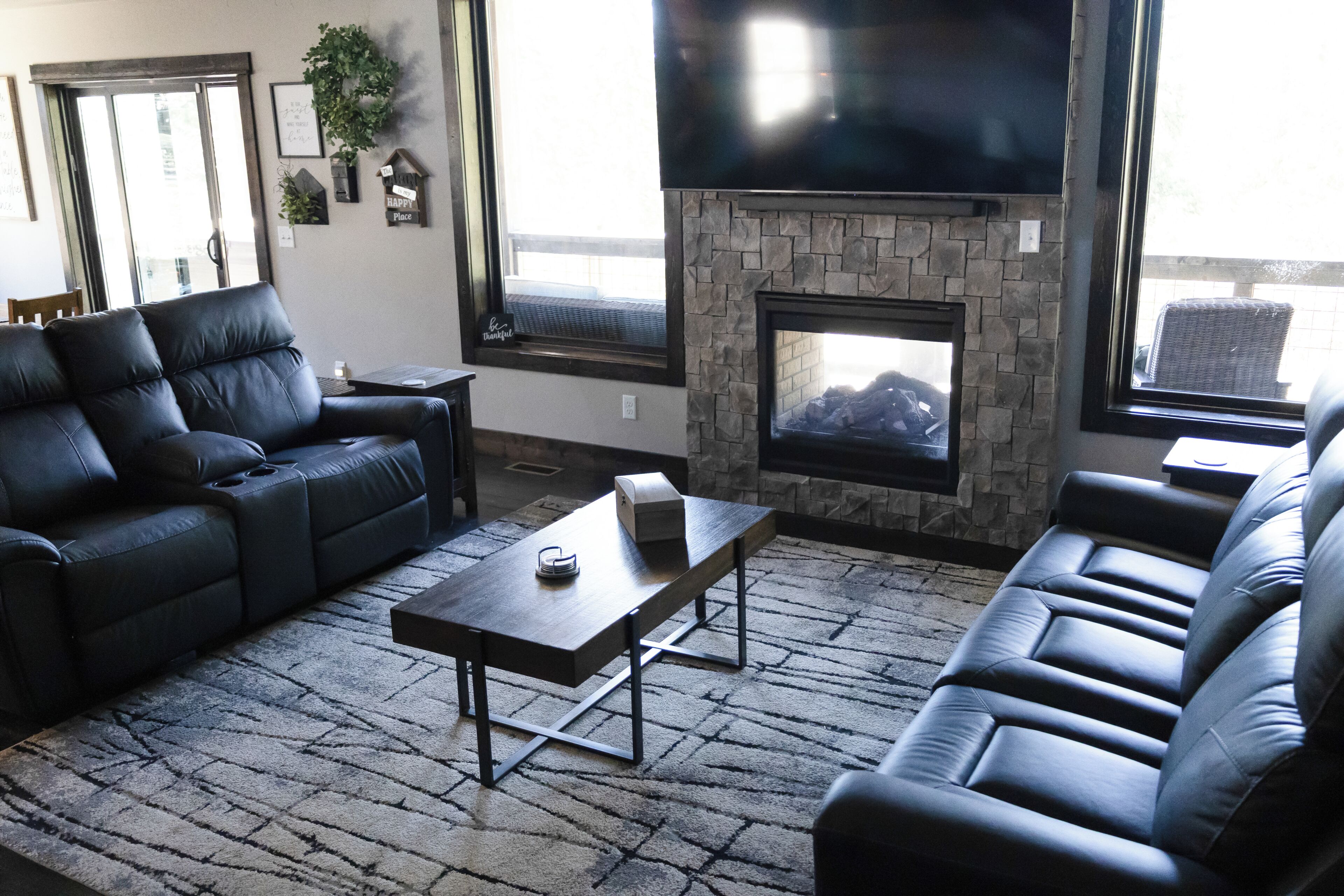 A living room with a stone fireplace, black leather sofas, and a flat-screen TV.
