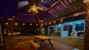 Un espace extérieur aménagé en patio, avec une table et des bancs en bois, un ventilateur de plafond et des guirlandes lumineuses.
