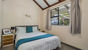 Ein Schlafzimmer mit Bett, Nachttisch, gerahmtem Bild, Lampe und einem Fenster mit Vorhängen.