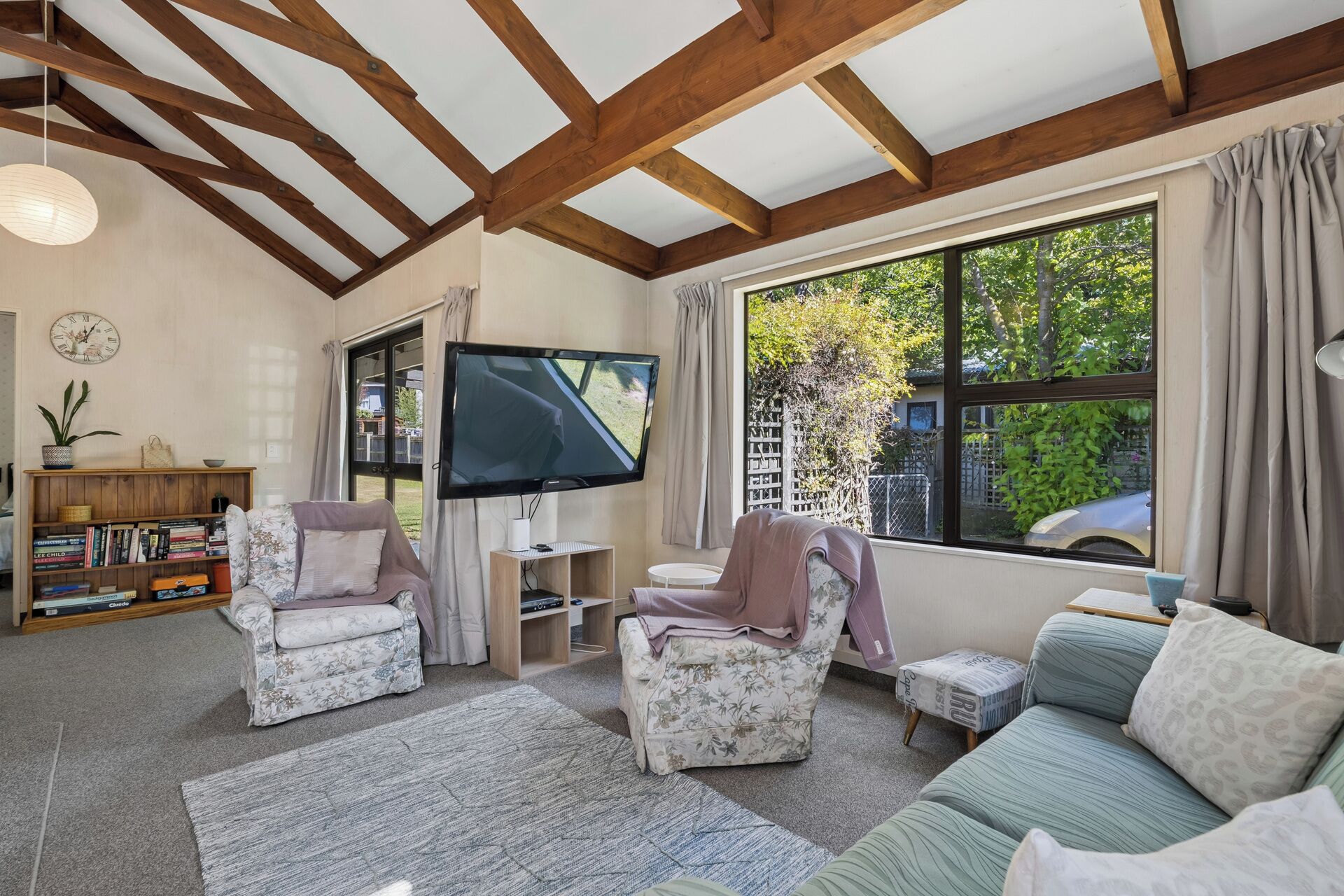 A cozy living room with a wooden ceiling, a flat-screen TV, a bookshelf, and a large window with a view of greenery.