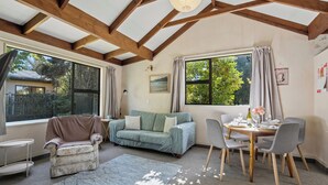 Ein gemütliches Zimmer mit Holzbalkendecke, einer Couch, einem Esstisch mit Stühlen und großen Fenstern mit Blick auf Grünflächen.
