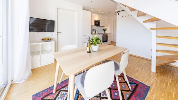 Una cocina moderna con armarios blancos, una mesa de comedor con sillas, un televisor montado sobre la encimera y una escalera con escalones de madera.