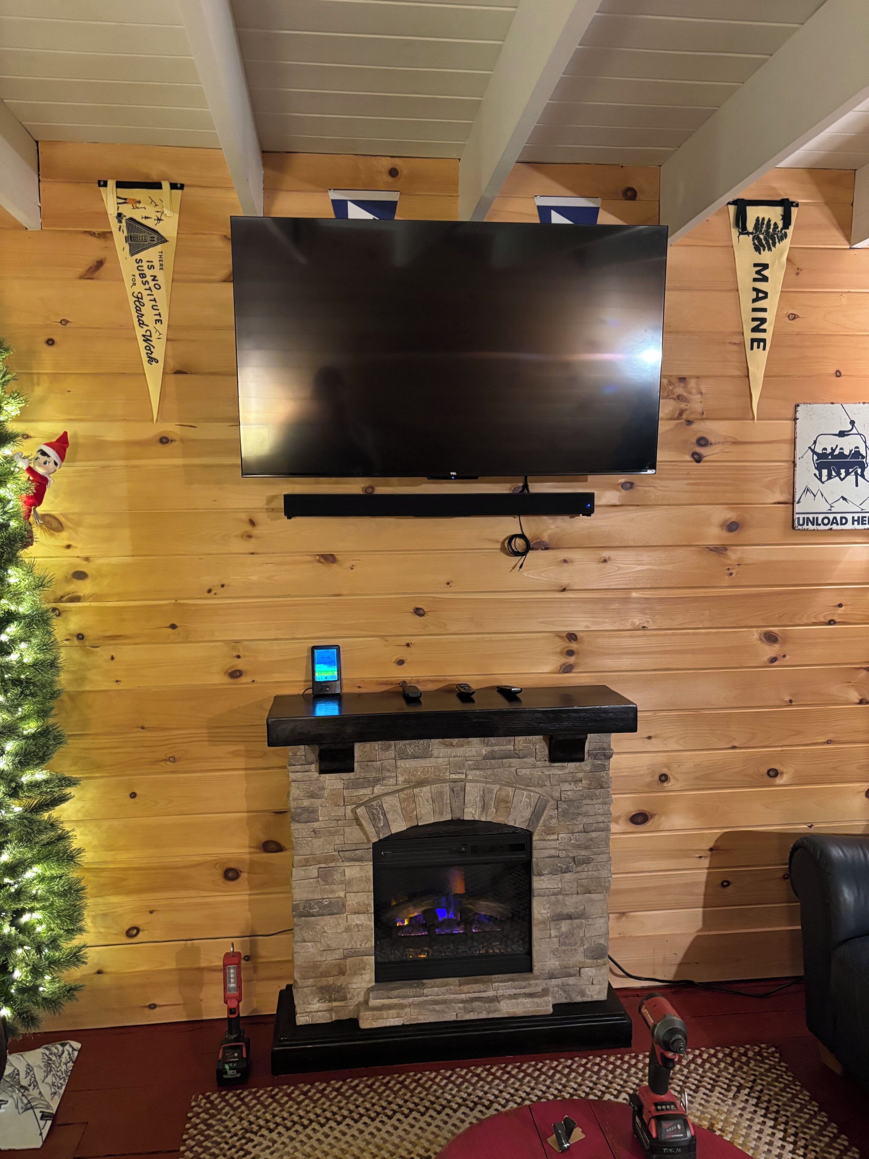 A living room with a flat-screen TV mounted on a wooden wall, a stone fireplace with a TV on top, and a Christmas tree with lights.