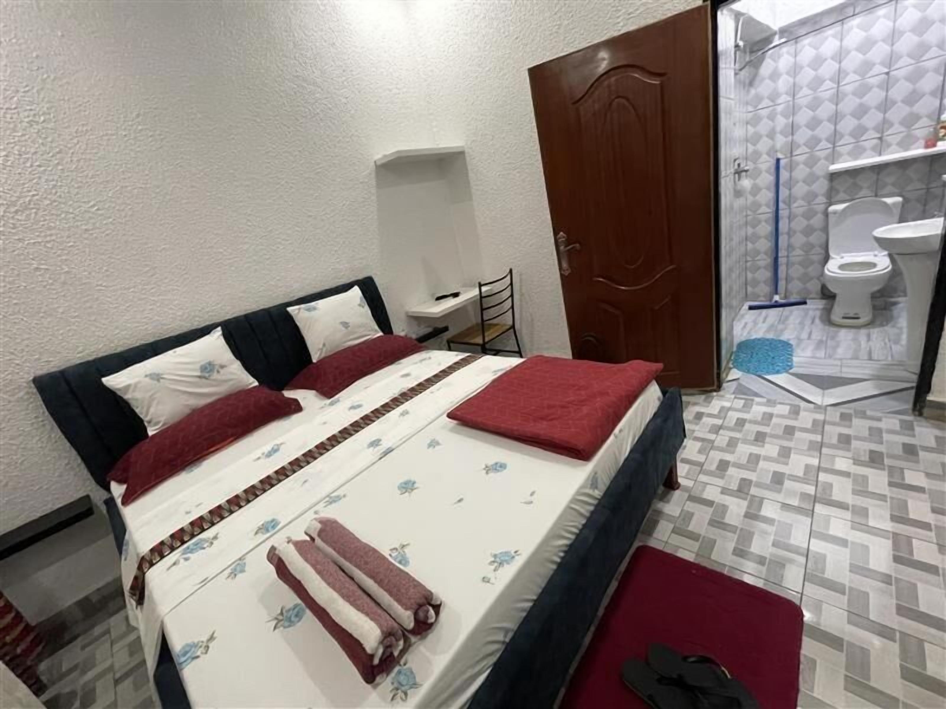 A neatly made bed with a white and red bedspread, a wooden headboard, and two red pillows. A wooden door and a bathroom with a white toilet and a blue bath mat are visible.