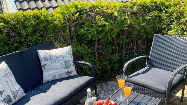 An outdoor seating area with a sofa and two chairs, a table with a bottle, a bowl of strawberries, a plate with watermelon, and a glass of orange juice.