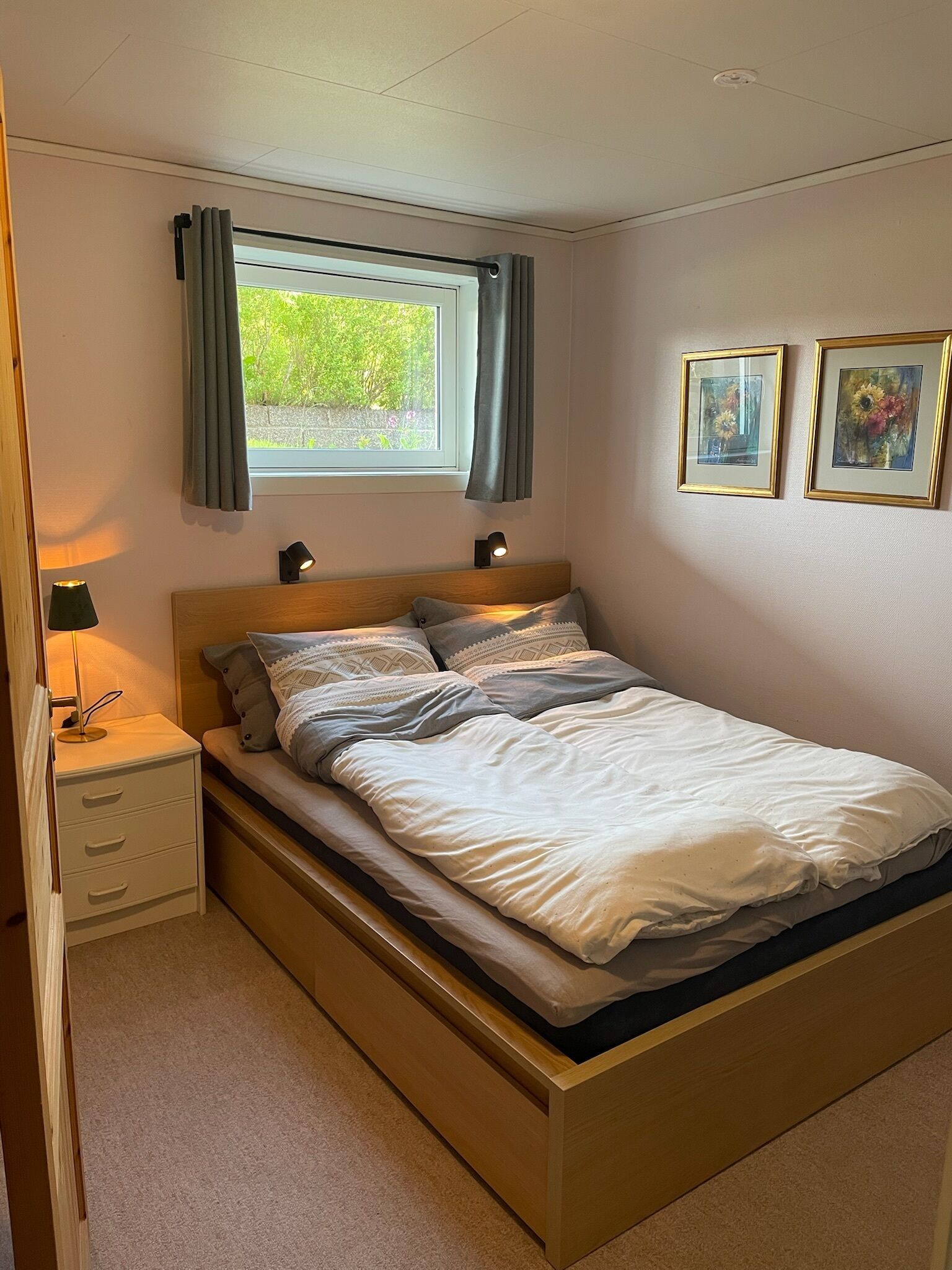 Een slaapkamer met een houten bed, een nachtkastje met een lamp, een raam met gordijnen, en twee ingelijste foto's aan de muur.