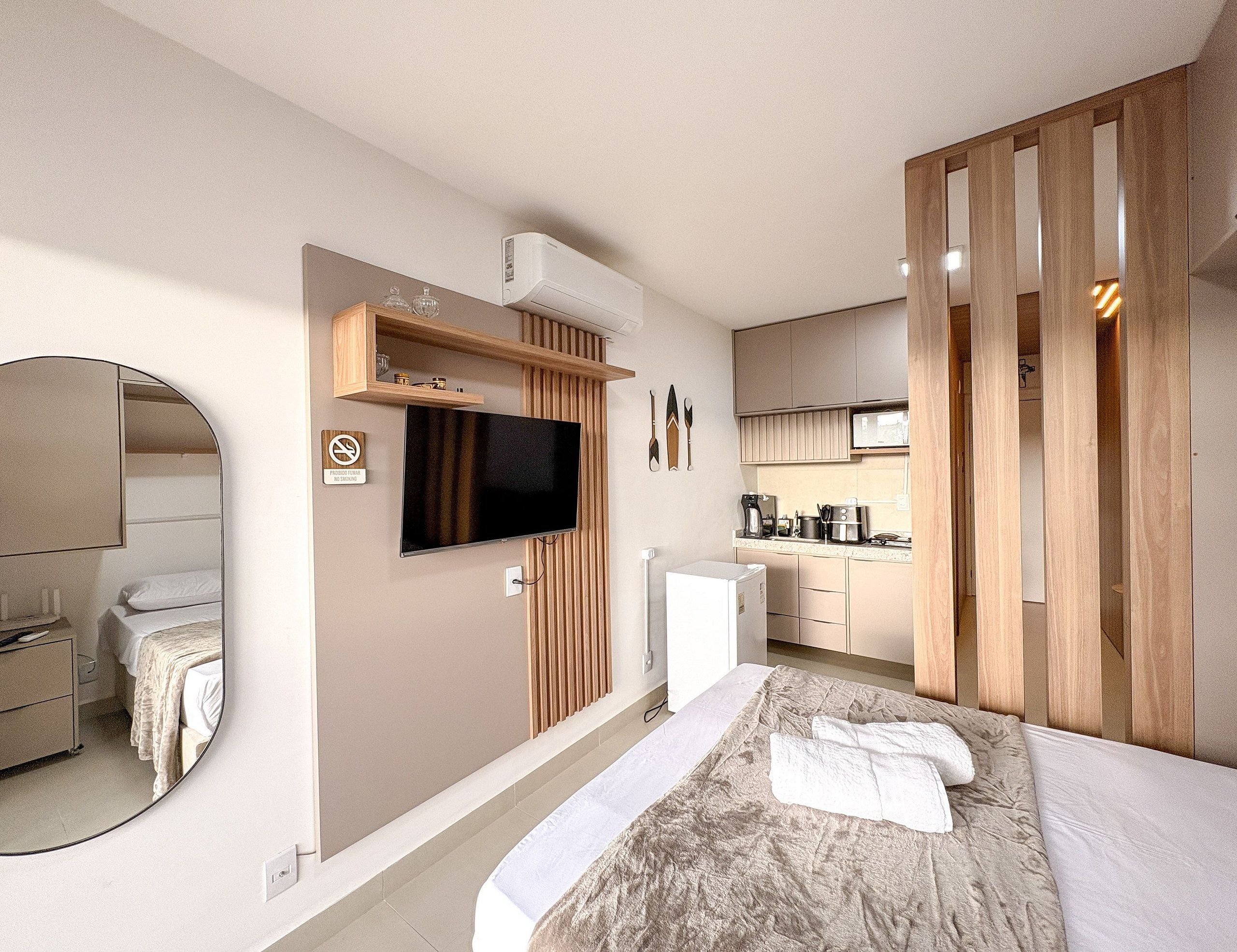 A modern hotel room with a bed, a TV, a kitchen area, and a mirror.