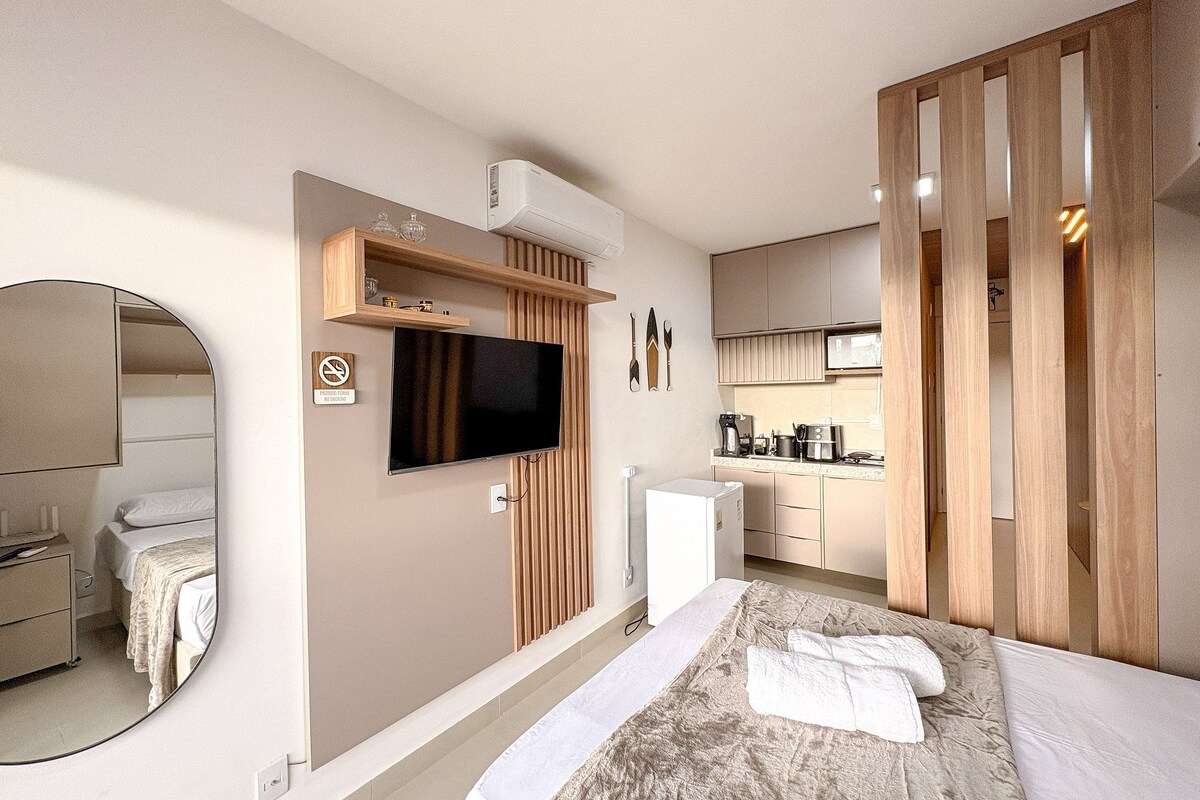 A modern hotel room with a bed, a TV, a kitchen area, and a mirror.