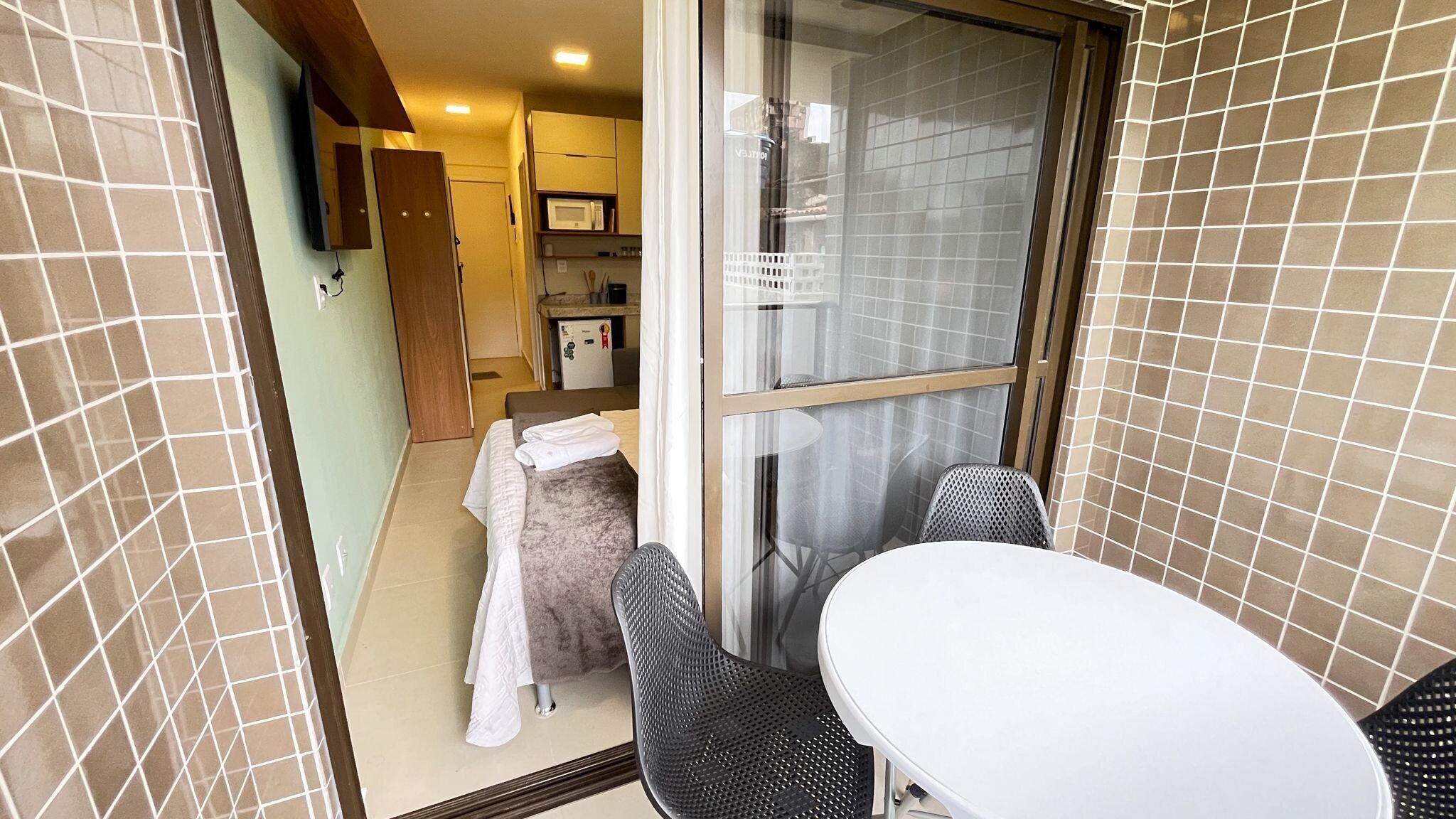 A small room with a sliding glass door leading to a balcony, a white table, two chairs, a bed with a white blanket, and a kitchenette with a microwave.