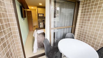 A small room with a sliding glass door leading to a balcony, a white table, two chairs, a bed with a white blanket, and a kitchenette with a microwave.