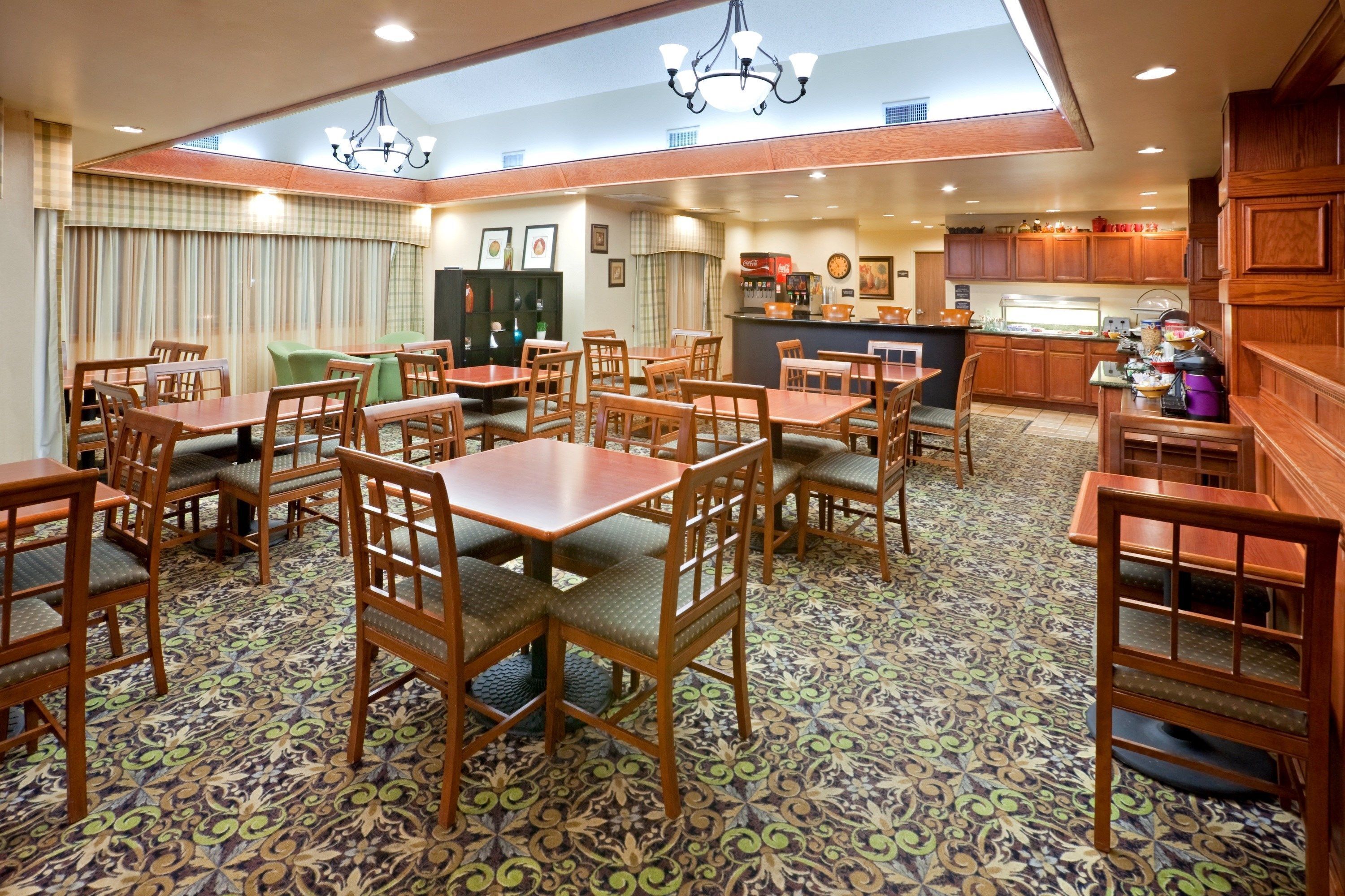 A dining area with wooden tables and chairs, a kitchen in the background, and patterned floor tiles.