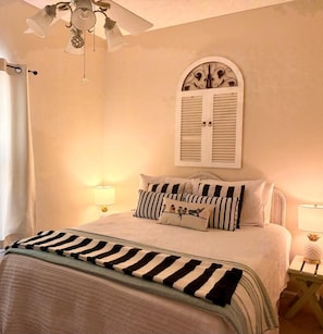 A bedroom with a bed, striped bedding, a decorative ceiling fan, and a wall-mounted arched window with shutters. -  2 Min Walk to the Beach - Sleeps 6 - 3BR Townhome with Private Outdoor Shower (Mexico Beach)