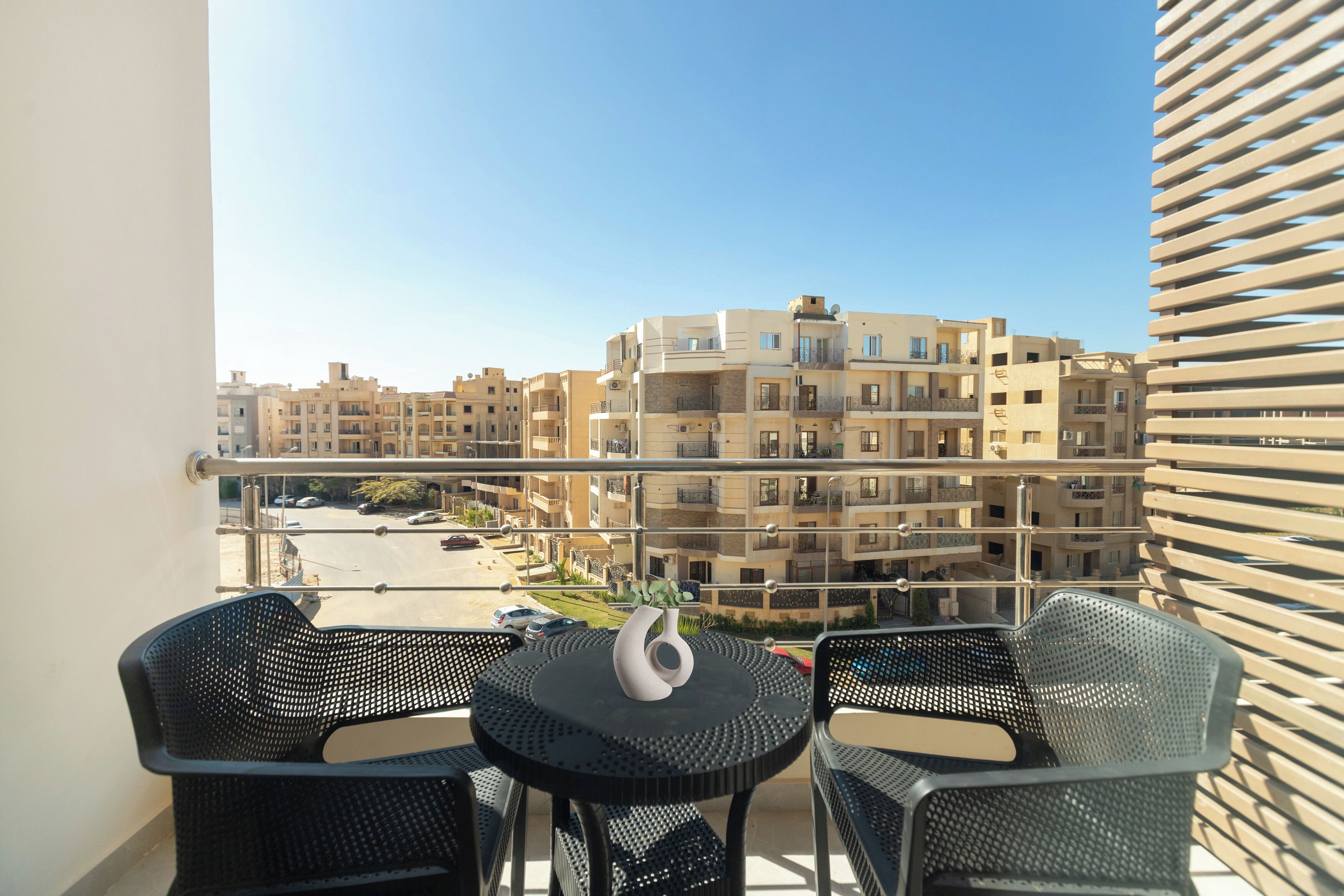 Uma varanda com duas cadeiras e uma mesa pequena, com vista para uma área residencial com vários edifícios de apartamentos.
