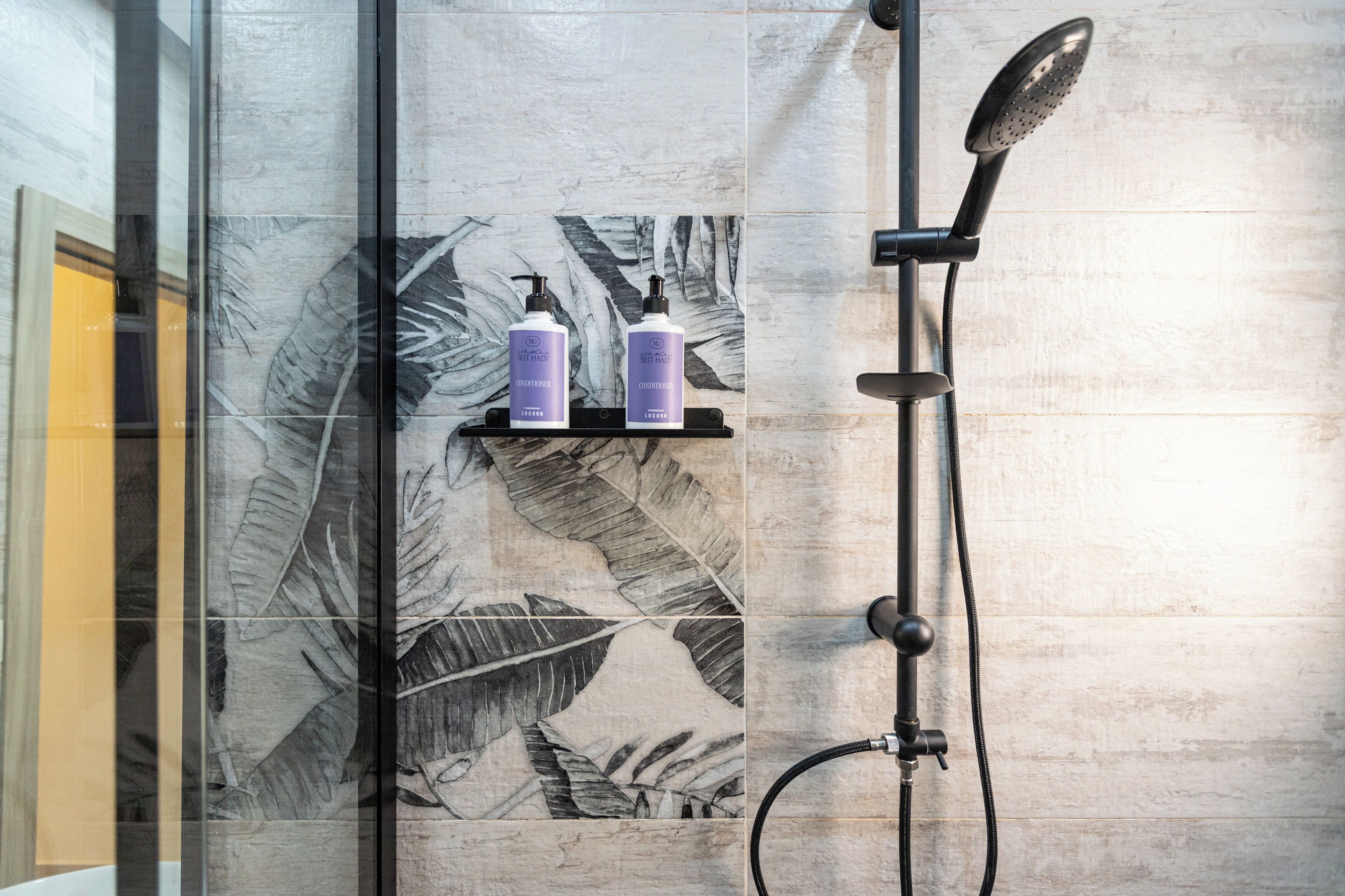A shower area with a glass door, a black showerhead, and two bottles of lotion on a shelf.