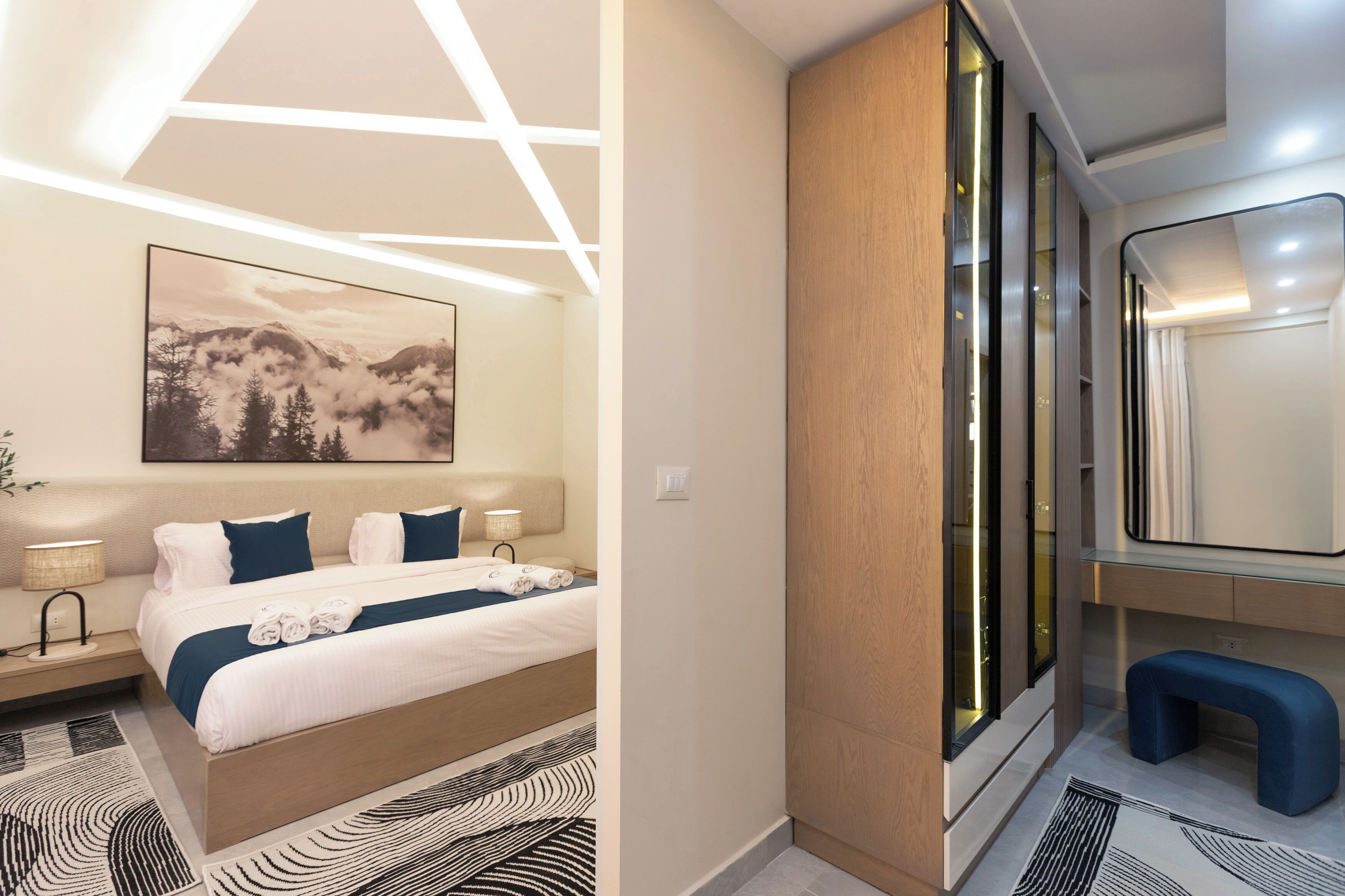 A modern hotel room with a bed, bedside lamps, a large window, and a bathroom with a mirror and bench.