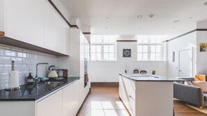 A modern kitchen with white cabinets, a black countertop, and a central island. There is a dining area with a gray sofa and a small table. - Stylish Historic Acton Apartment (London)