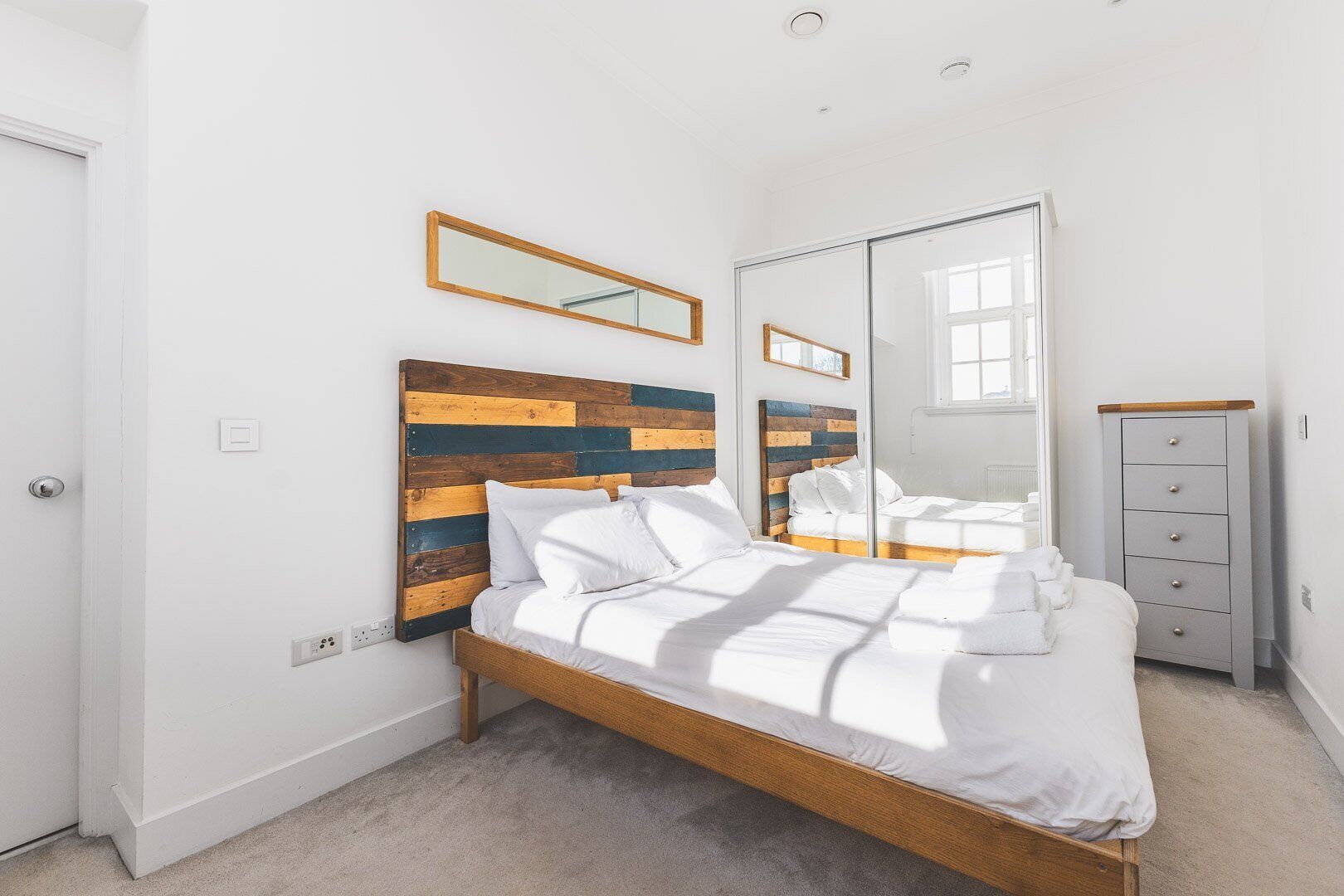 A bedroom with a wooden headboard, a large mirror, a wardrobe, and a bed with white bedding.