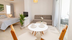 A modern hotel room with a bed, a sofa, a dining table with chairs, a TV, a ceiling fan, and a large window.