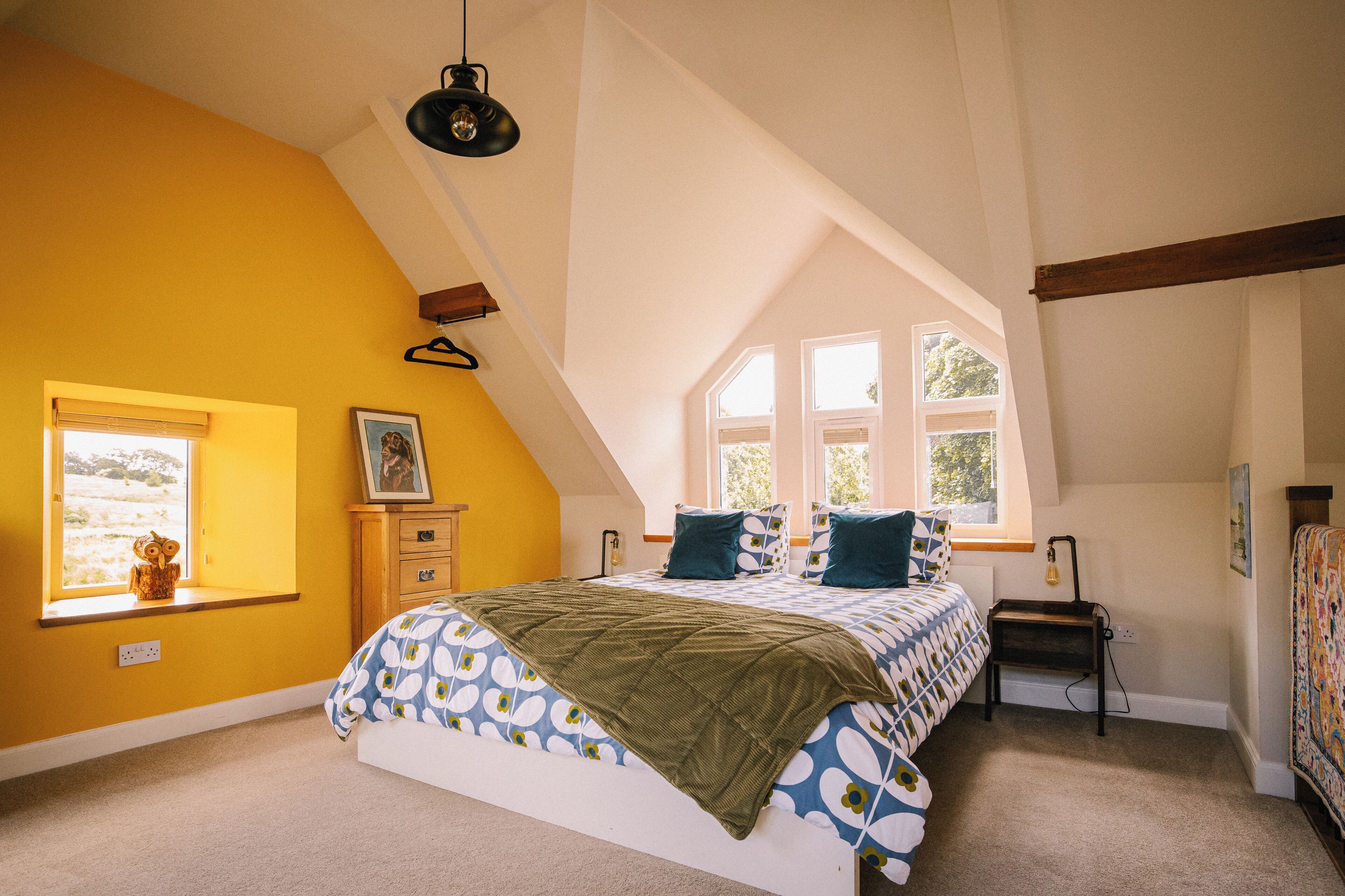 A bedroom with a sloped ceiling, a bed with patterned bedding, a small bedside table, and a window with a view of the outdoors.