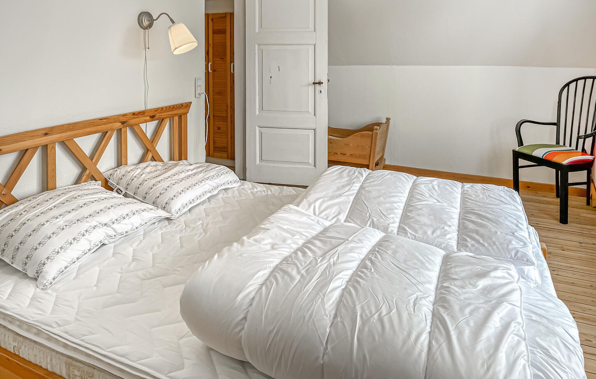 A bedroom with a wooden bed frame, white bedding, a bedside lamp, a wooden chair, and a door.
