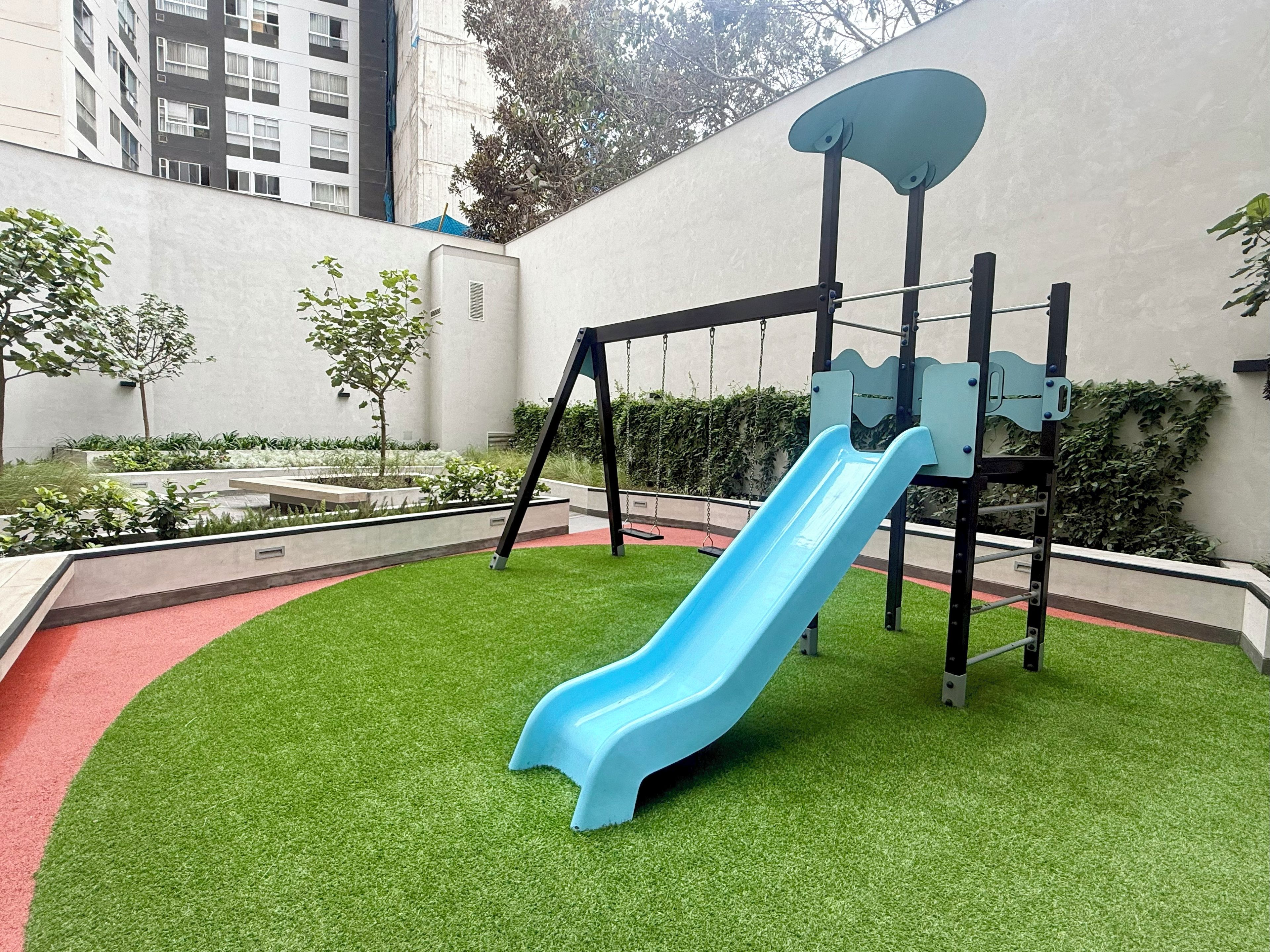 A playground with a blue slide and swings, surrounded by artificial grass and greenery.