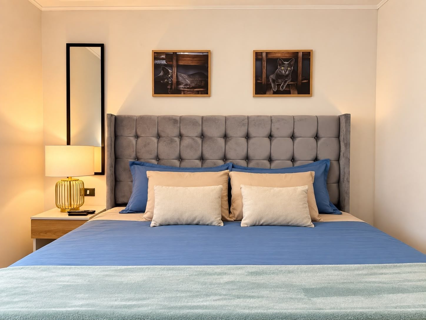 A bedroom with a tufted headboard, a bed with blue and beige bedding, two pillows, a bedside table with a lamp, and two framed pictures on the wall.