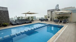 Una piscina al terrat amb gandules i ombrelles, envoltada de plantes en testos i edificis moderns.