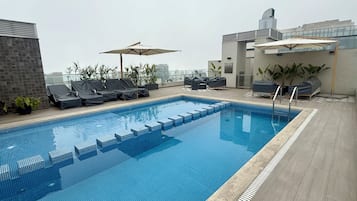 A rooftop swimming pool with lounge chairs and umbrellas, surrounded by potted plants and modern buildings.